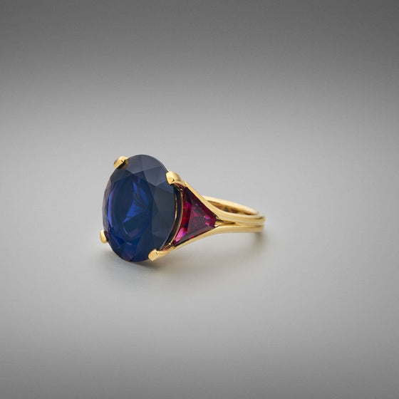 A BUNDA 'Bundova' Iolite and Rubellite Ring in 18ct yellow gold. The ring is set with a large round brilliant iolite in a four claw 18ct yellow gold setting. Two trapezoid cut rubellite tourmalines are bezel set to the shoulders. The shank is an 18ct yellow gold double wire wrap.  Characteristics of Iolite - 1 = 18.75cts  Characteristics of Rubellite Tourmaline - 2 = 3.10cts  Weight of Ring = 13.30 grams