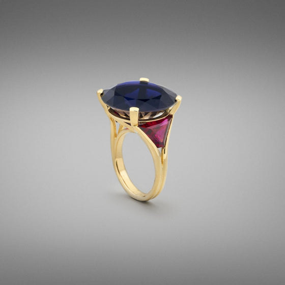 A BUNDA 'Bundova' Iolite and Rubellite Ring in 18ct yellow gold. The ring is set with a large round brilliant iolite in a four claw 18ct yellow gold setting. Two trapezoid cut rubellite tourmalines are bezel set to the shoulders. The shank is an 18ct yellow gold double wire wrap.  Characteristics of Iolite - 1 = 18.75cts  Characteristics of Rubellite Tourmaline - 2 = 3.10cts  Weight of Ring = 13.30 grams