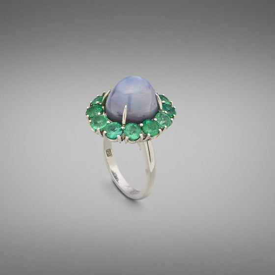 A BUNDA 'Cluster' ring made in platinum, centre set with a high domed blue star sapphire in a flour claw platinum setting. The centre stone is surrounded by a cluster of round brilliant cut emeralds in four claw settings.  Characteristics of Star Sapphire: 1 = 18.15ct  Characteristics of Emeralds: 12 = 3.35cts  Ring is stamped 'BUNDA' Total weight: 14.70 grams